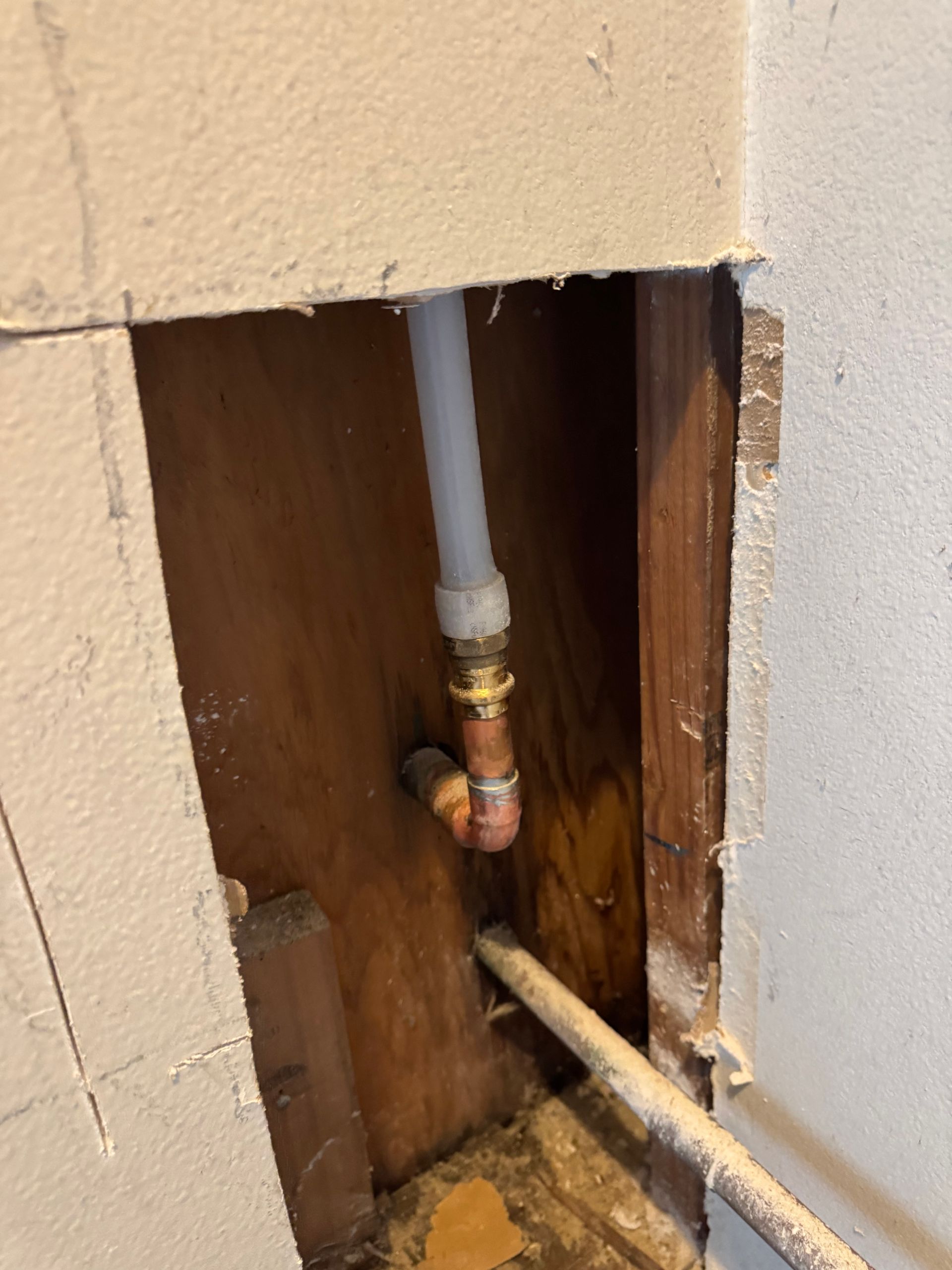 Pipes in wall cavity; copper elbow joint connects to PEX pipe, and metal pipe. Brown wood and beige wall.