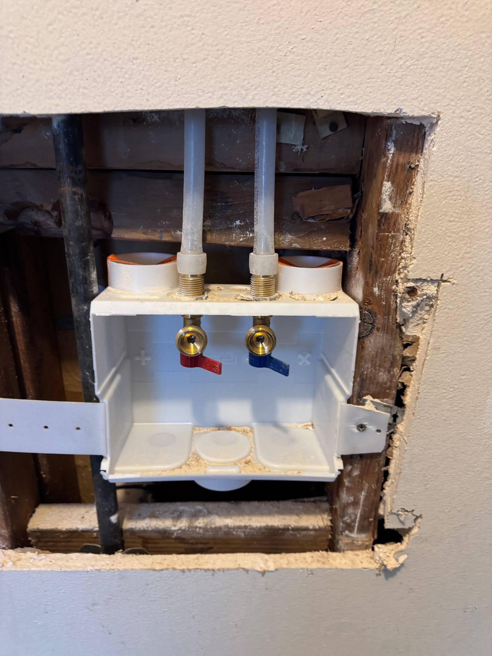 White laundry box with red and blue water valves and PEX pipes within a wall opening.