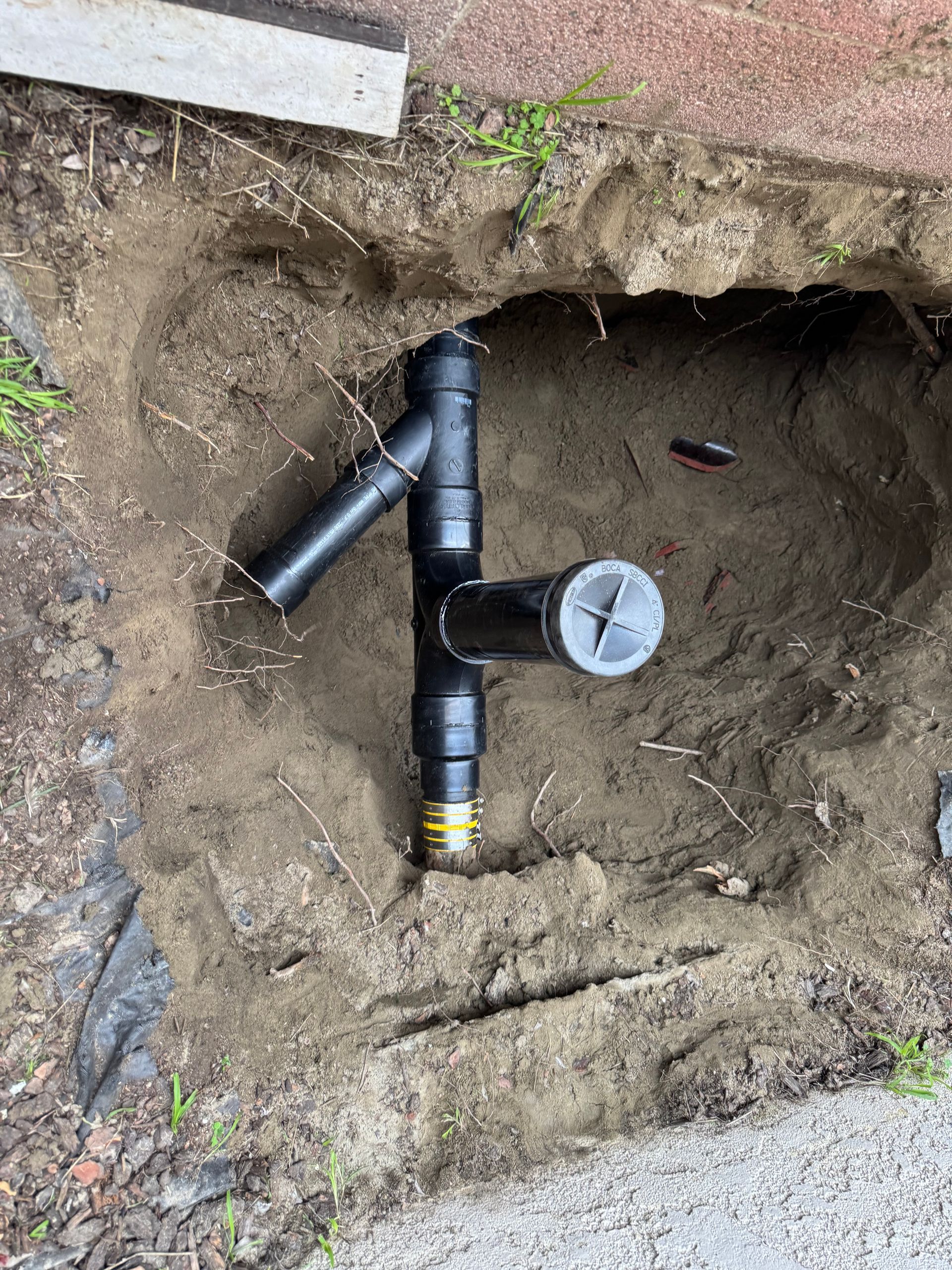 Black PVC drain pipes in a dirt hole, connected with a T-fitting and capped outlet.