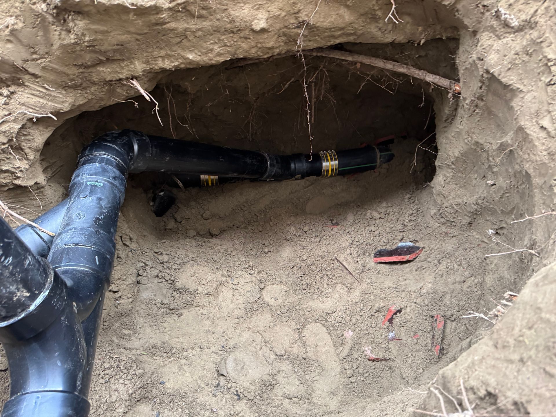 Black pipes in a hole dug in the ground with dirt walls. Some wire is also visible.