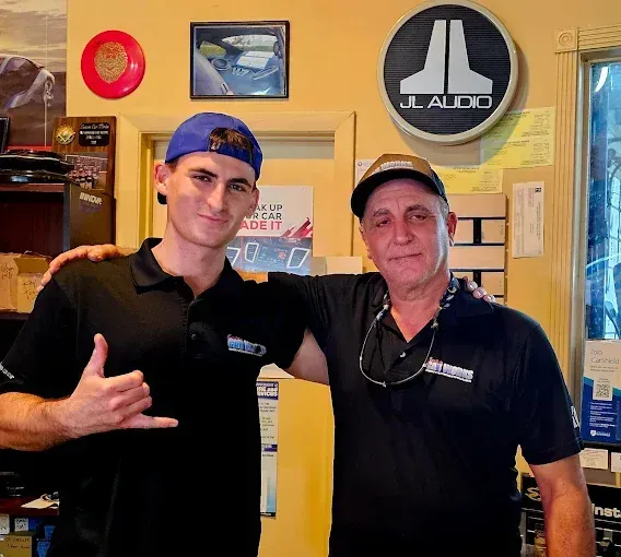 Two men in black shirts posing indoors, one giving a hand gesture, with wall signs behind them.