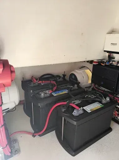 Two large black batteries with red cables in a utility corner, beside equipment and a pink wheel.