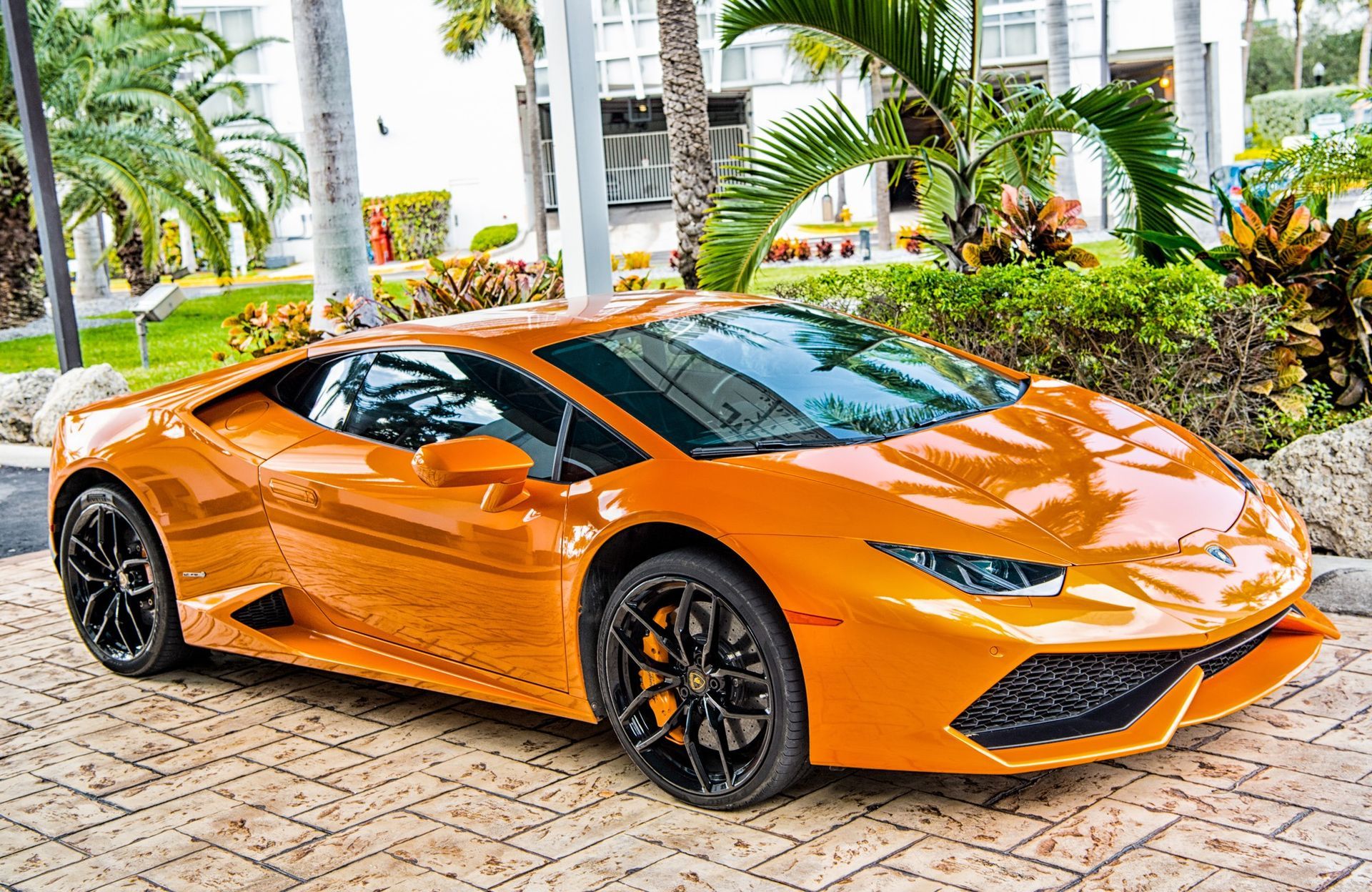 A bright orange Lamborghini sports car parked on a stone-paved driveway in front of a building with tropical landscaping.