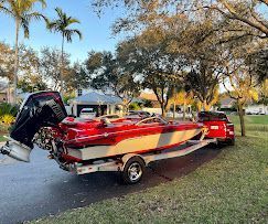 Red and white speedboat on a trailer beside a pickup in a grassy park under trees.