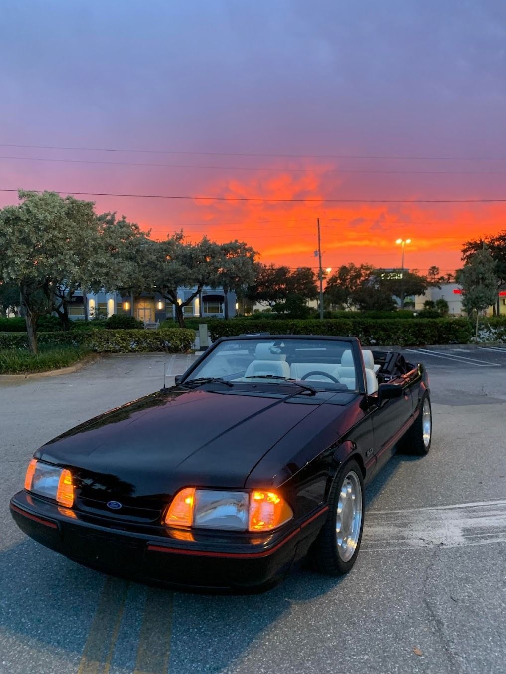 A black Ford Mustang convertible parked in an empty parking lot against a vibrant orange and purple sunset sky.