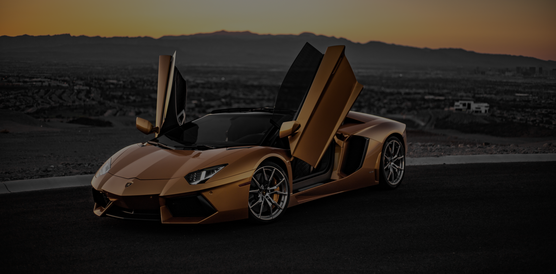 A bronze Lamborghini Aventador with its scissor doors open, parked on a paved road during sunset.