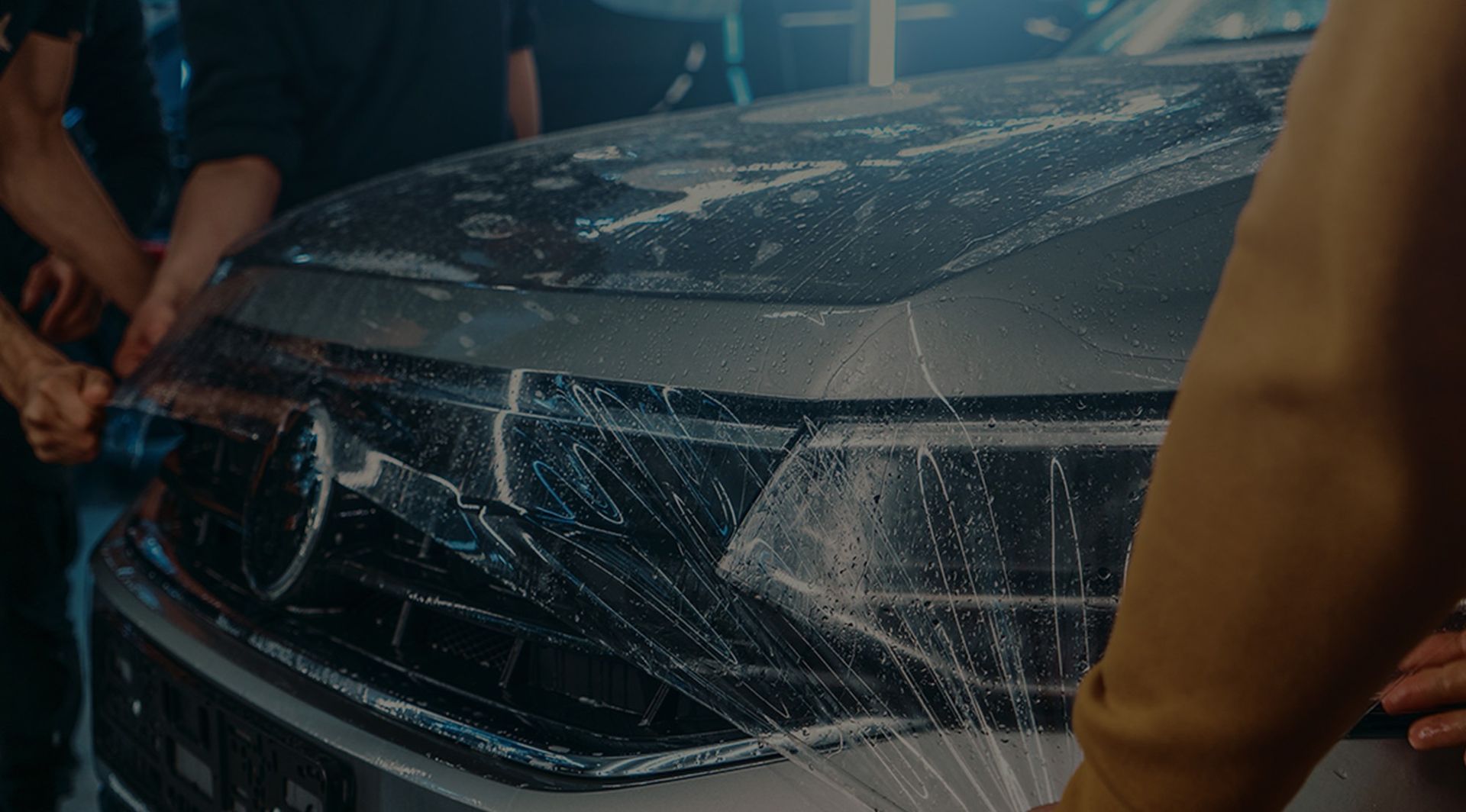 Several people carefully apply a transparent protective film onto the front grill and hood of a silver car.