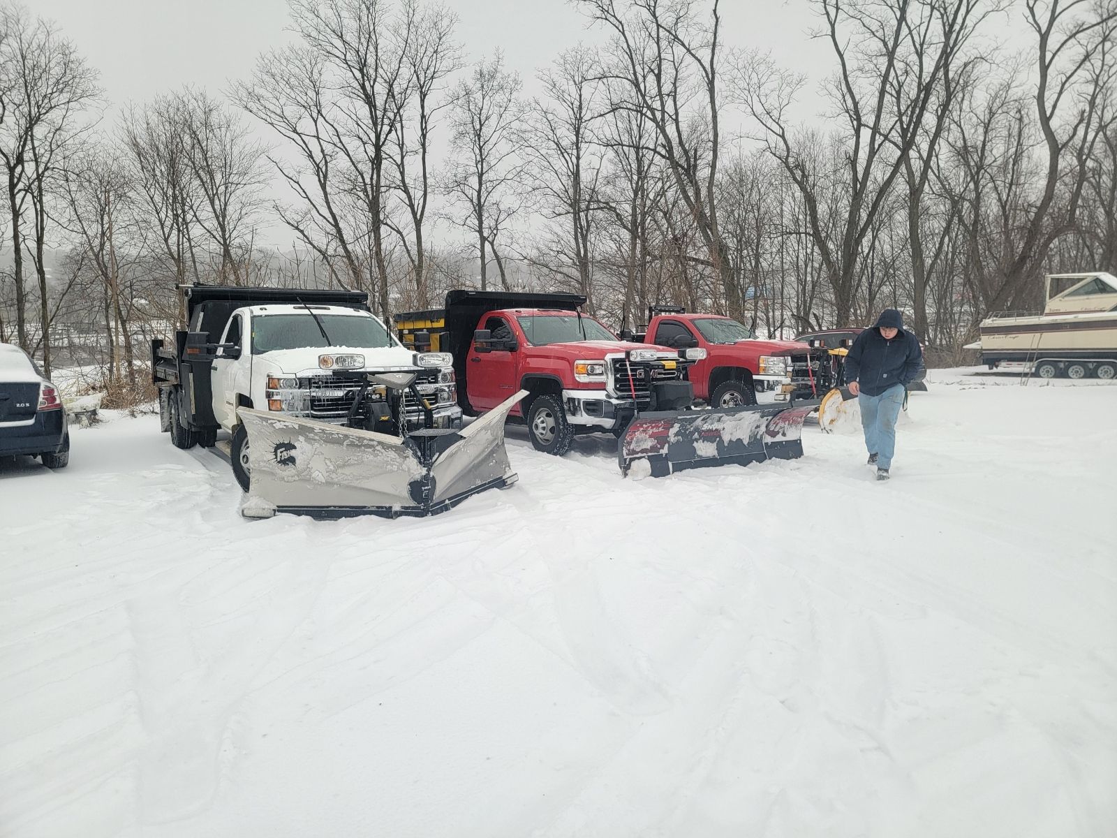 Snow-covered lot with two plow trucks, a person walking, and bare trees.