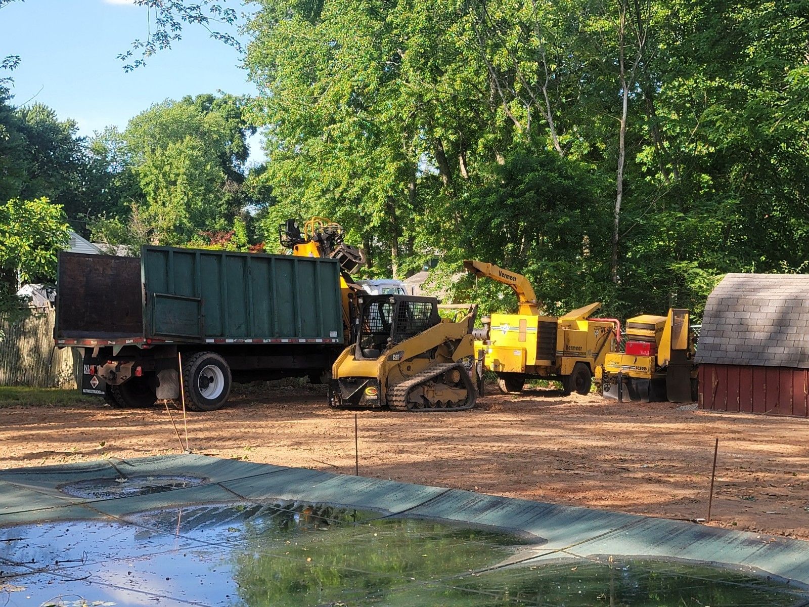 Truck and machinery loading wood chips in a yard. Green truck, yellow chipper, and shed.