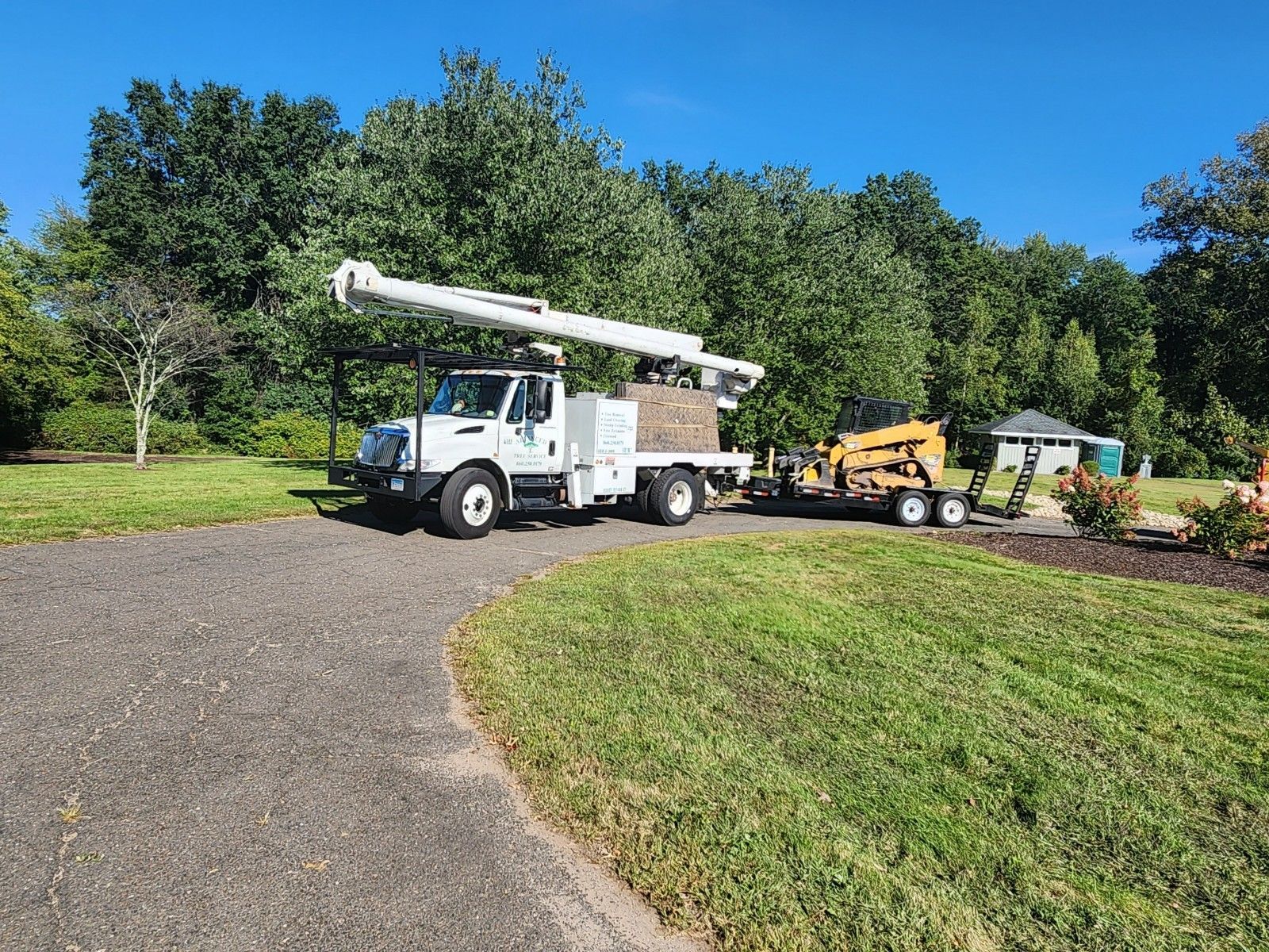 White utility truck with an extended arm, towing a wood chipper, parked on gravel next to grass.