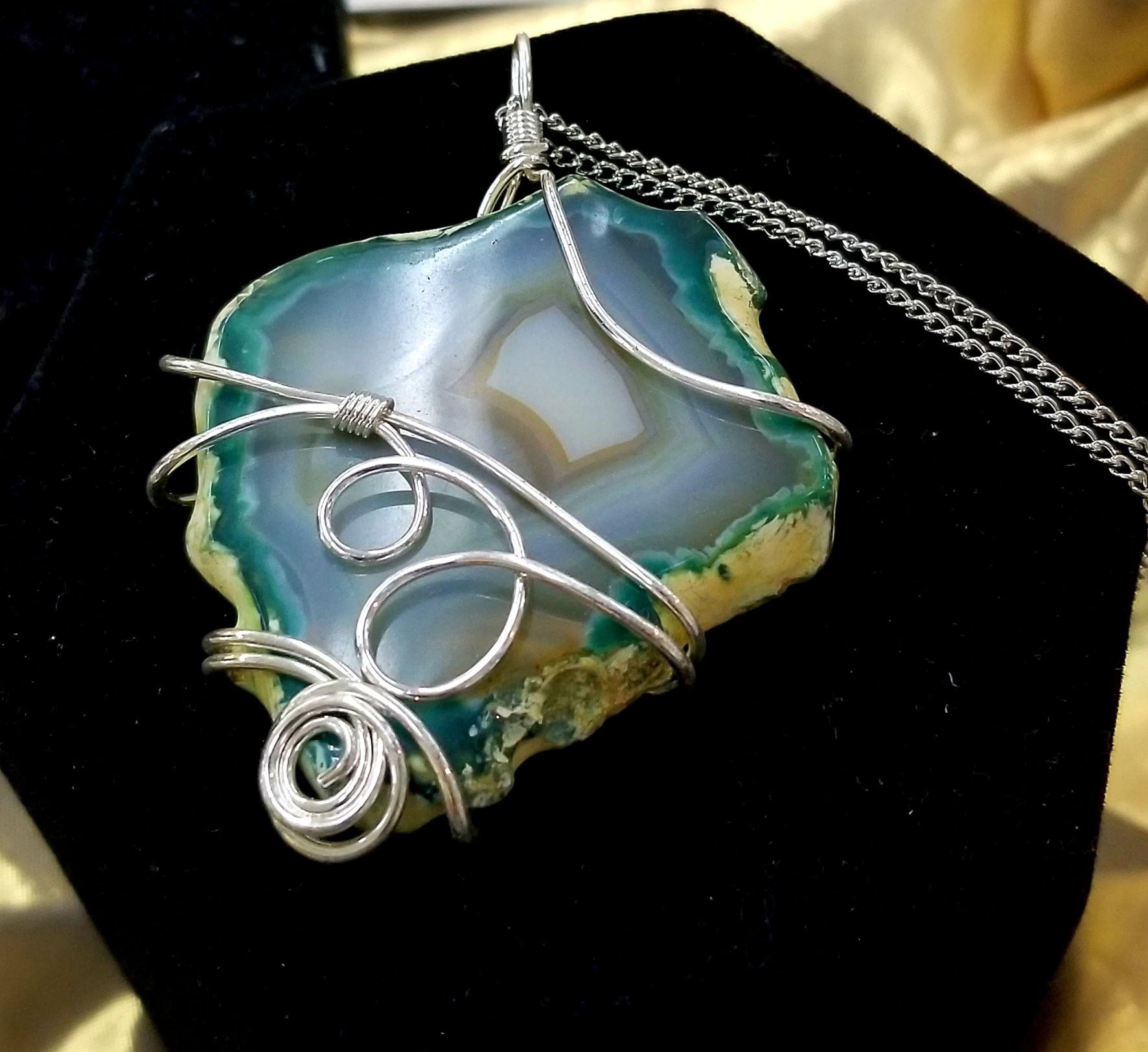 A wire-wrapped agate pendant with a silver chain, resting on a black velvet surface. The stone is blue, white, and green.