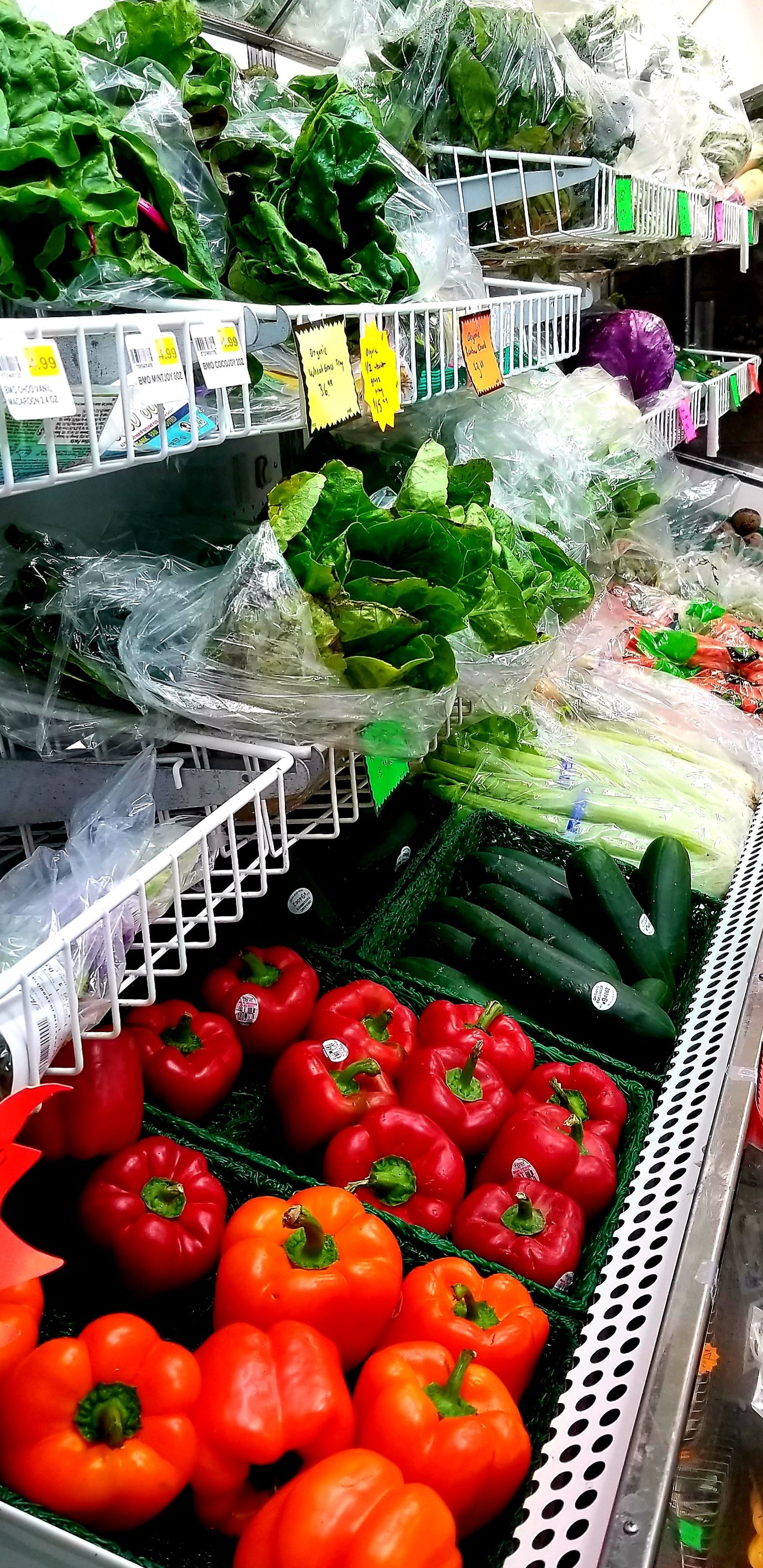 Produce display in a grocery store, with red bell peppers, cucumbers, lettuce, and other vegetables.