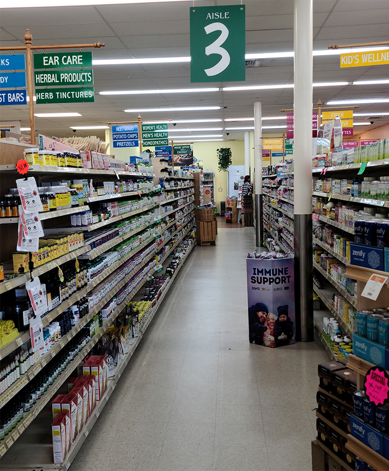 Grocery store aisle with shelves of products, green signs overhead.