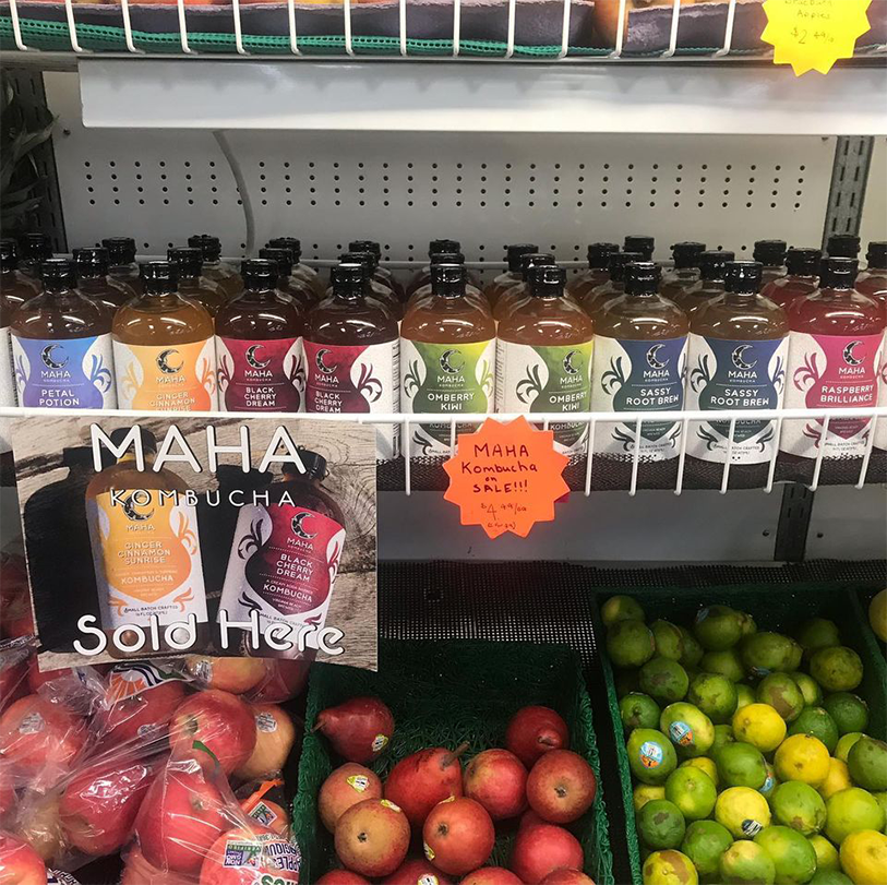 Bottles of Maha Kombucha on a shelf above a produce section of fruit.