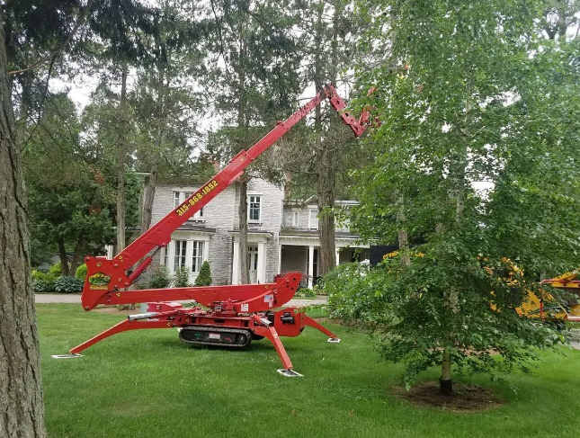 Red tracked crane boom parked on grass in front of a white house, partly hidden by trees.
