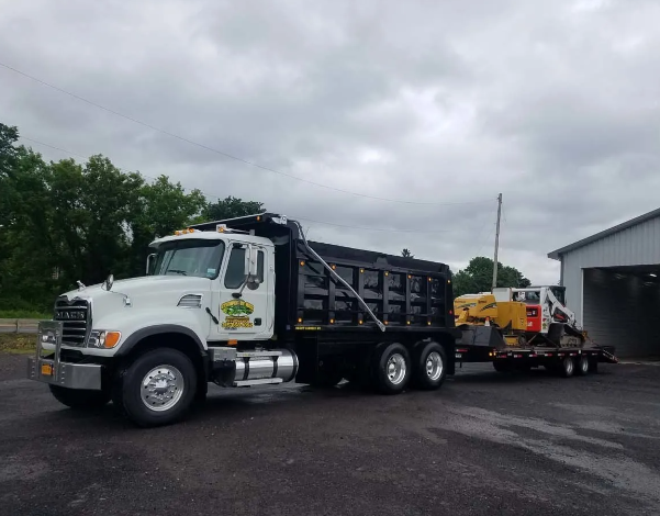 White dump truck hauling heavy equipment on a trailer beside a warehouse on a cloudy day