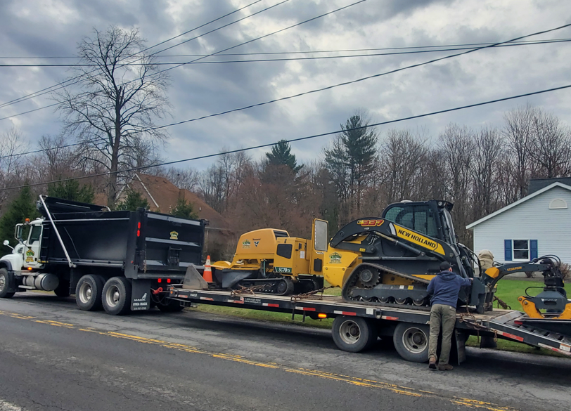 Black flatbed trailer hauling yellow construction equipment on a roadside, with a dump truck and worker nearby.
