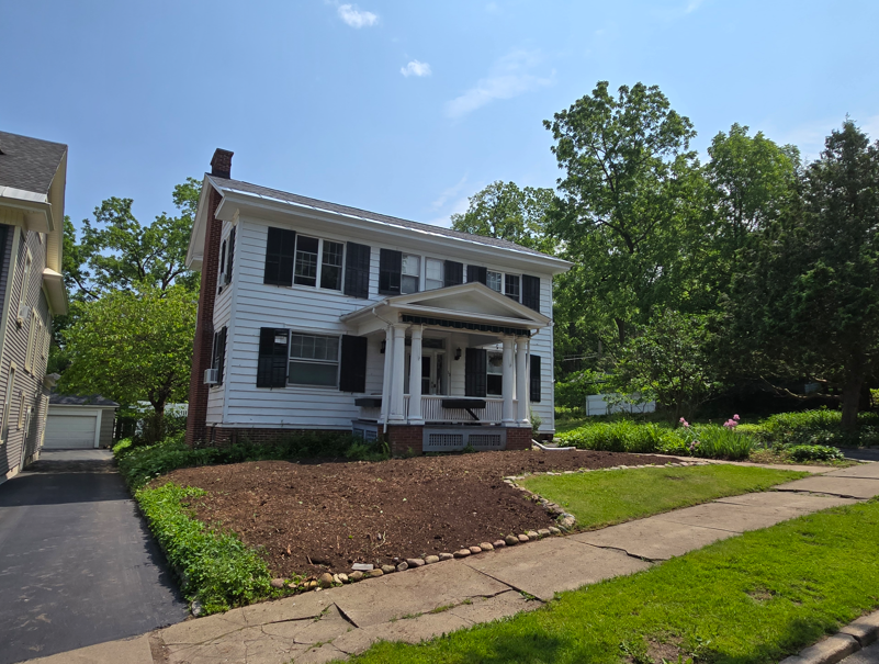 Two-story white house with front porch, black shutters, and a landscaped yard on a sunny day