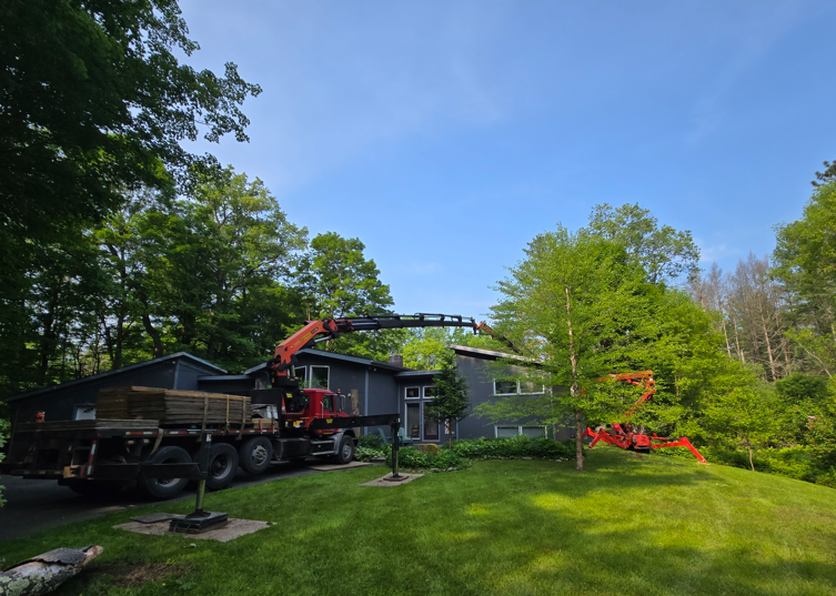 Tree removal crew using a crane to lift logs beside a house on a grassy yard