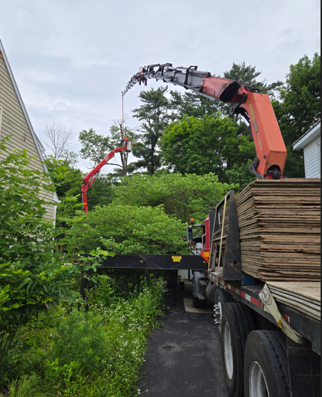 Orange crane unloading stacked lumber from a truck in a narrow driveway