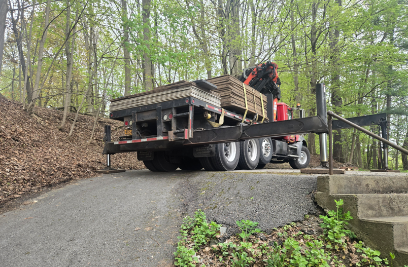 Flatbed truck loaded with lumber slowly navigating a narrow wooded driveway beside concrete steps.