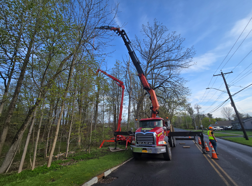 Utility truck with red crane trimming trees along a roadside, with workers on the shoulder.