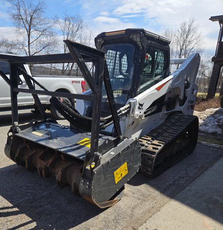 Compact tracked loader with grapple attachment parked outdoors on pavement.