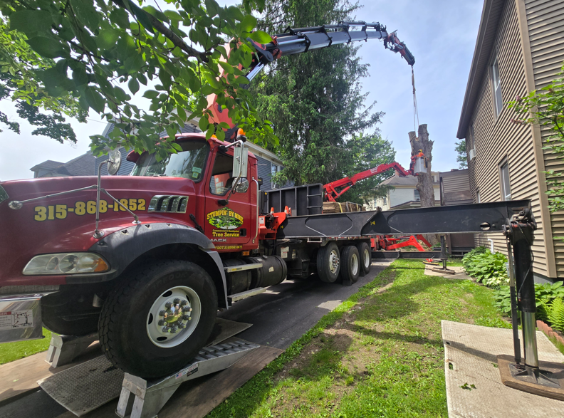 Tow truck hoists a car in a narrow driveway beside a brick house and green lawn.