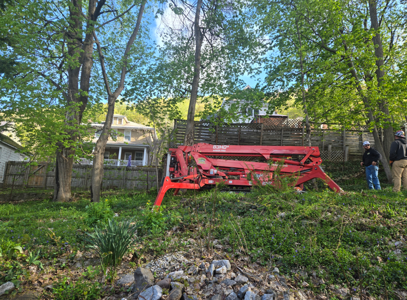 Red farm machine on a grassy hillside with trees and a wooden fence in the background.