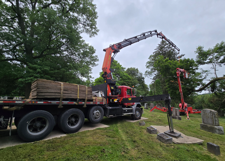 Crane truck lifting a bundled load beside a grassy roadside with trees and stone markers