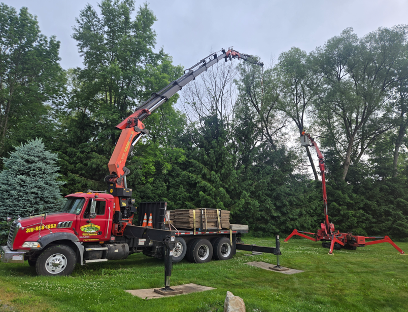 Red truck with orange crane lifting a tree trunk in a grassy yard with trees