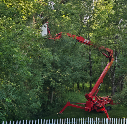 Red boom lift with extended arm working among tall green trees beside a white fence.