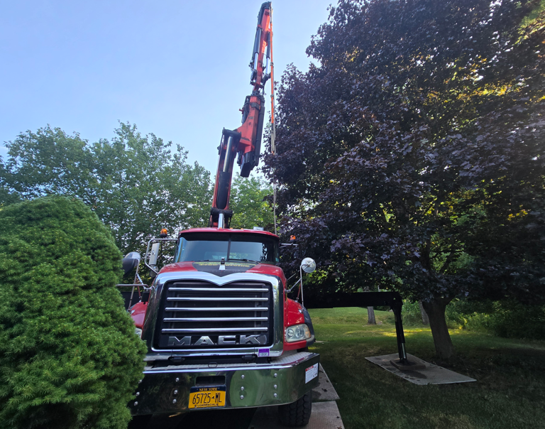 Red Mack truck with crane parked in a leafy yard beside a large purple tree.