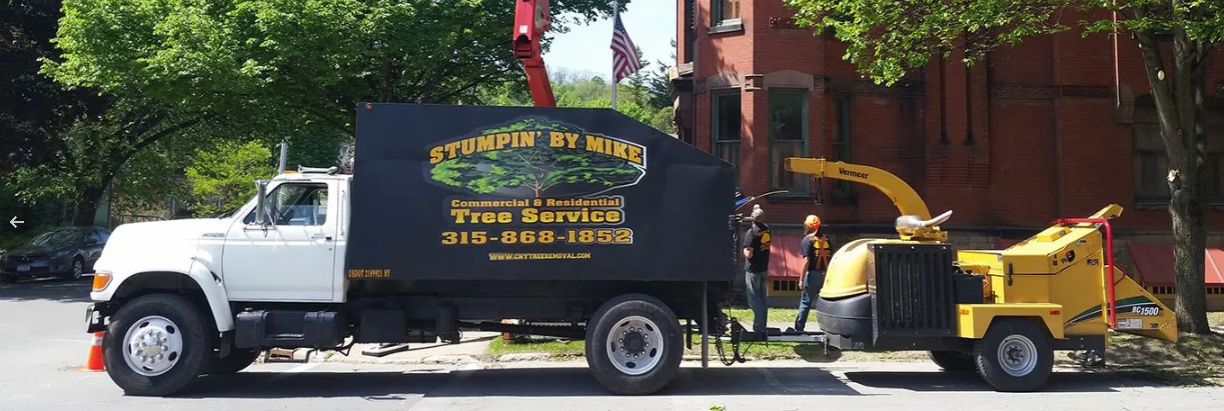 White truck towing a yellow tow machine on a city street beside a brick building