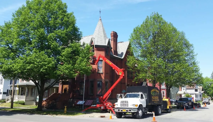 Red lift working on a red Victorian house with a pointed tower, trees and parked truck on a sunny street
