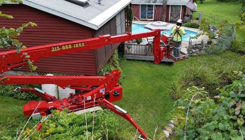 Red telescopic boom lift parked beside a house, with workers trimming overgrown greenery in a yard.