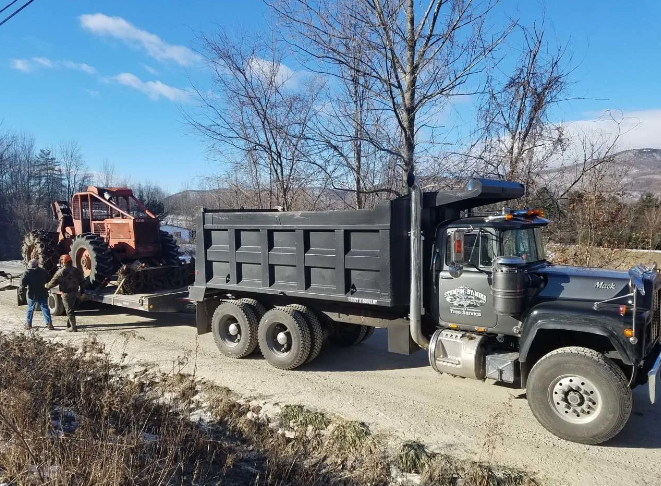 Dump truck towing a red farm machine along a rural dirt road on a sunny day