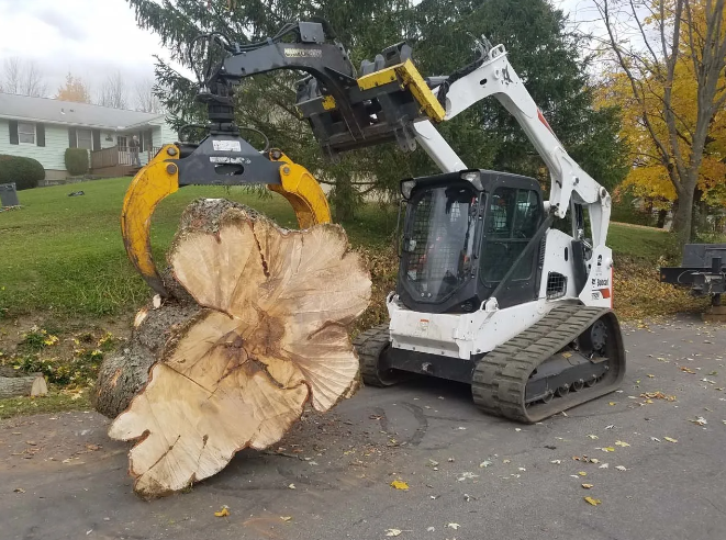 White skid-steer loader lifting a giant tree stump on a residential street with a grappling attachment