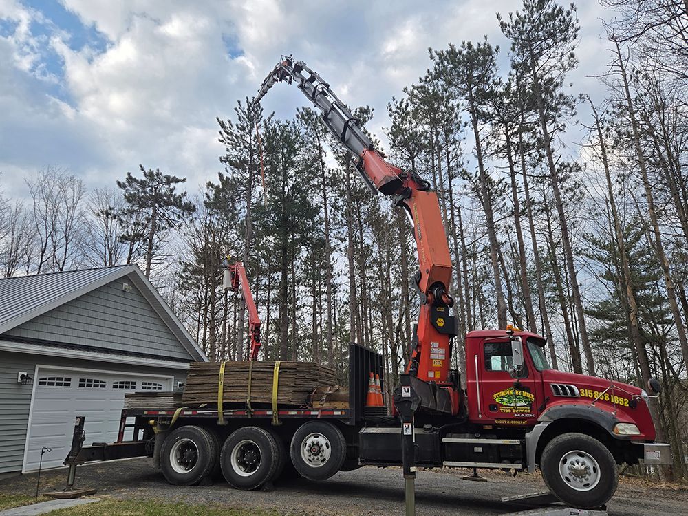 Red crane truck loading logs beside a garage in a wooded driveway
