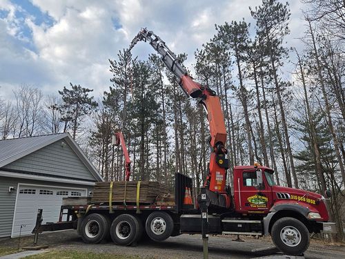 Red crane truck loading logs beside a garage in a wooded driveway