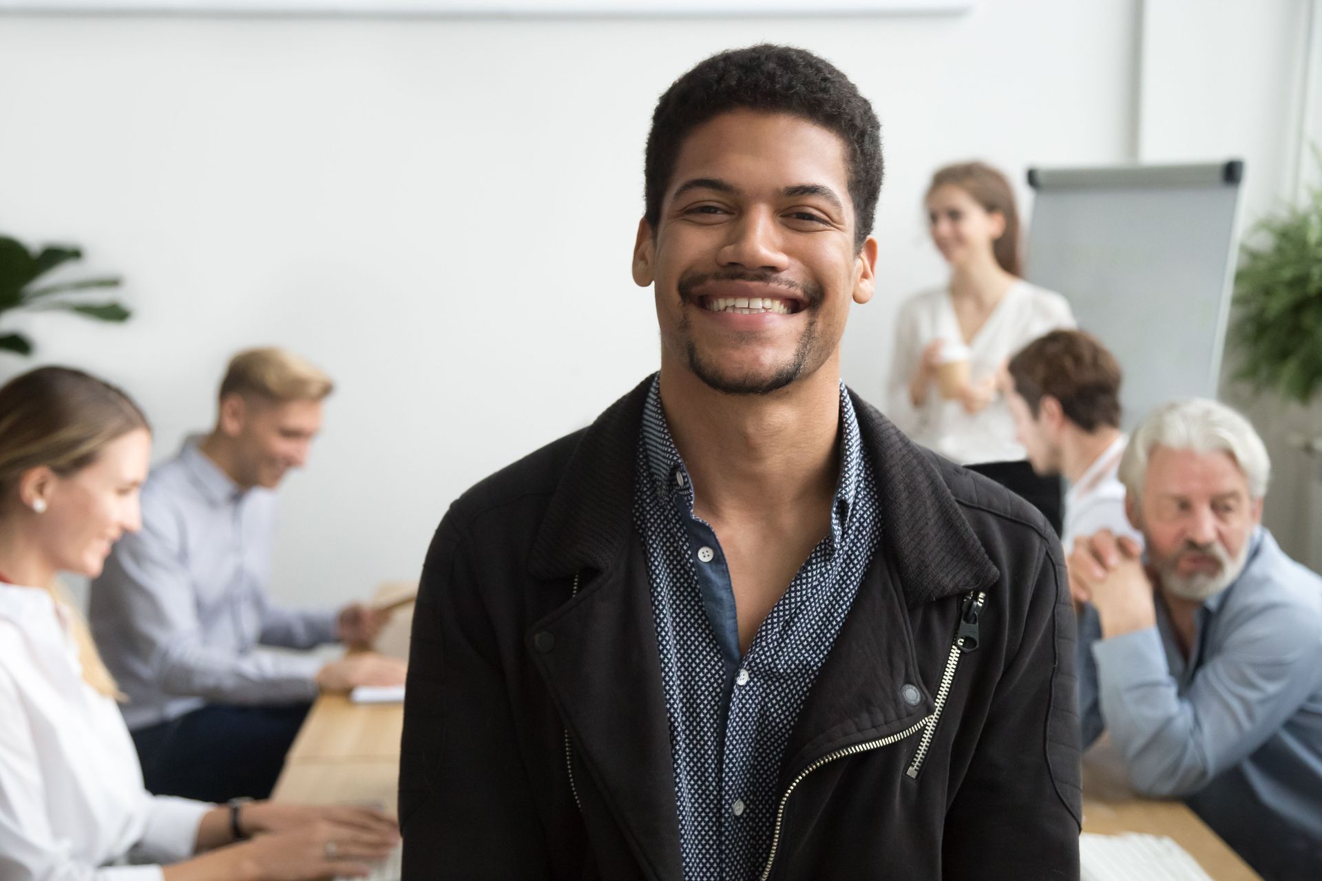 A smiling person in a black jacket stands in a blurred office setting with colleagues working in the background.