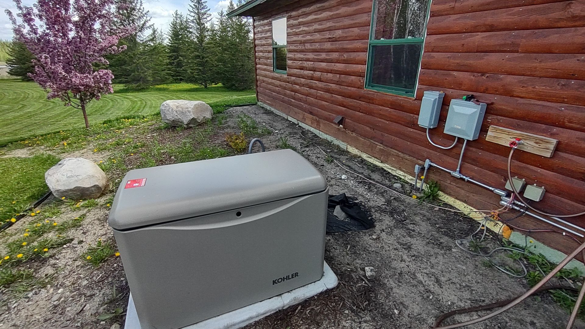 A gray standby generator sits outside a log cabin, with electrical boxes and conduit mounted on the exterior wall.