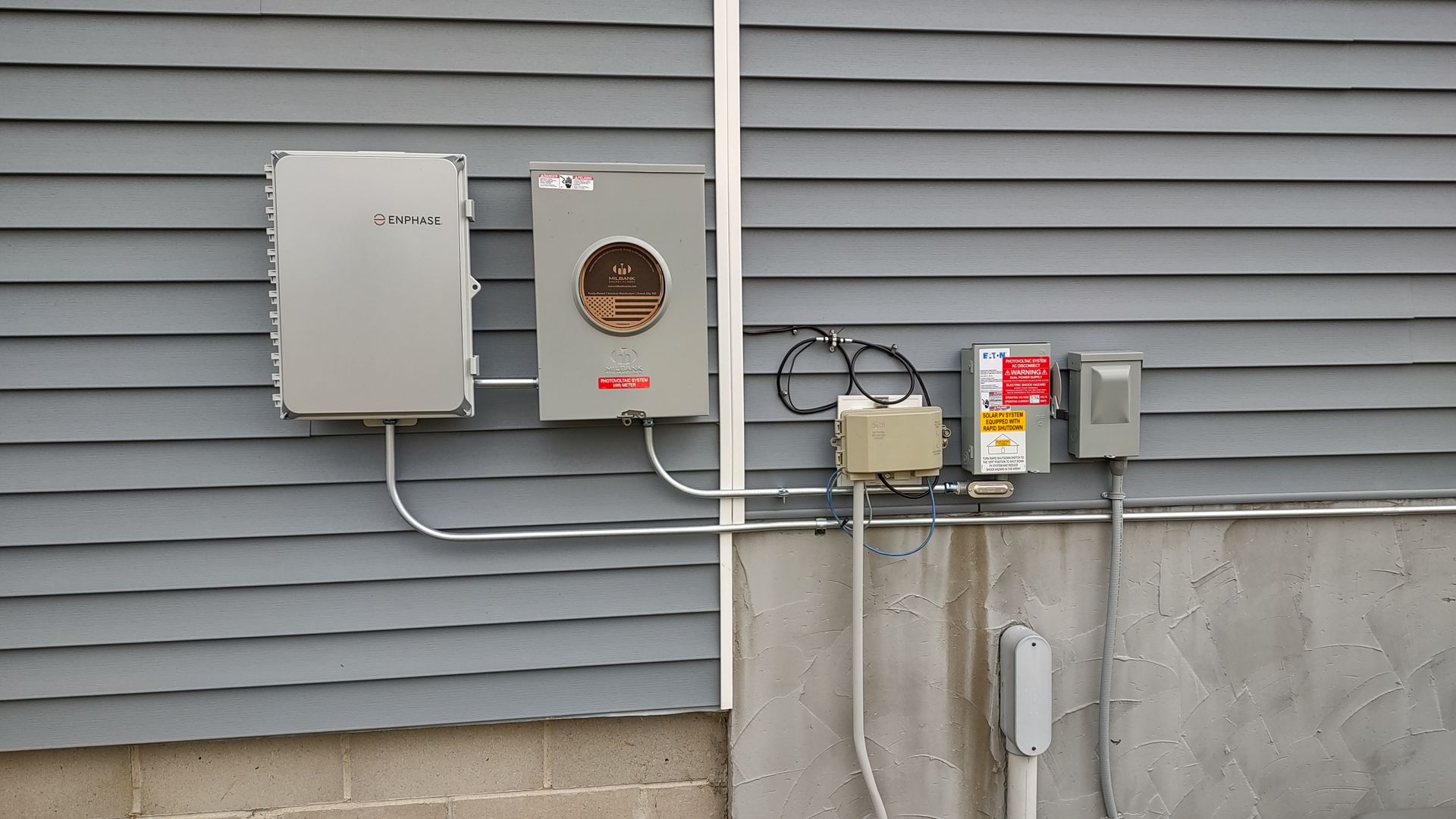Electrical equipment, including a meter box and conduit, mounted on a gray vinyl-sided exterior wall.