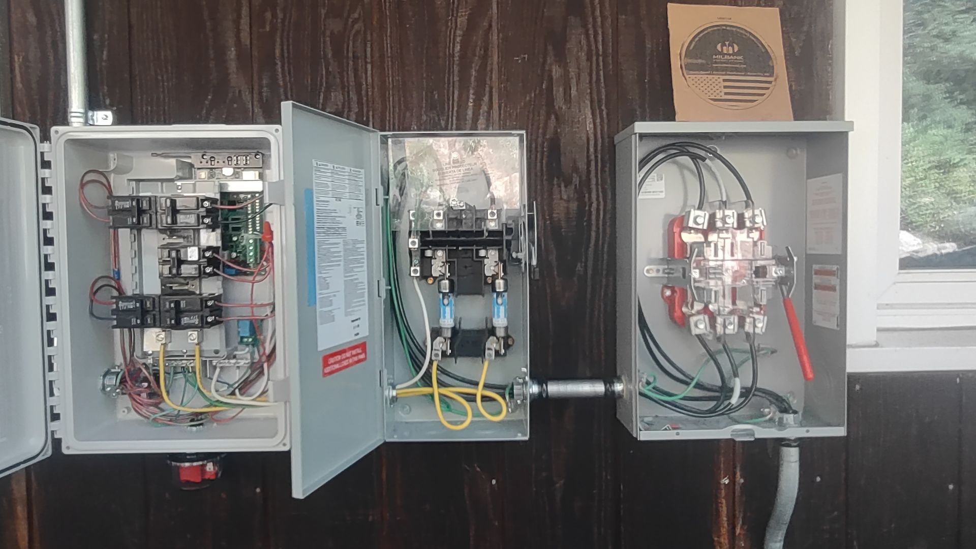 Three open, wall-mounted gray electrical enclosures connected by conduit on a dark wood-paneled wall.