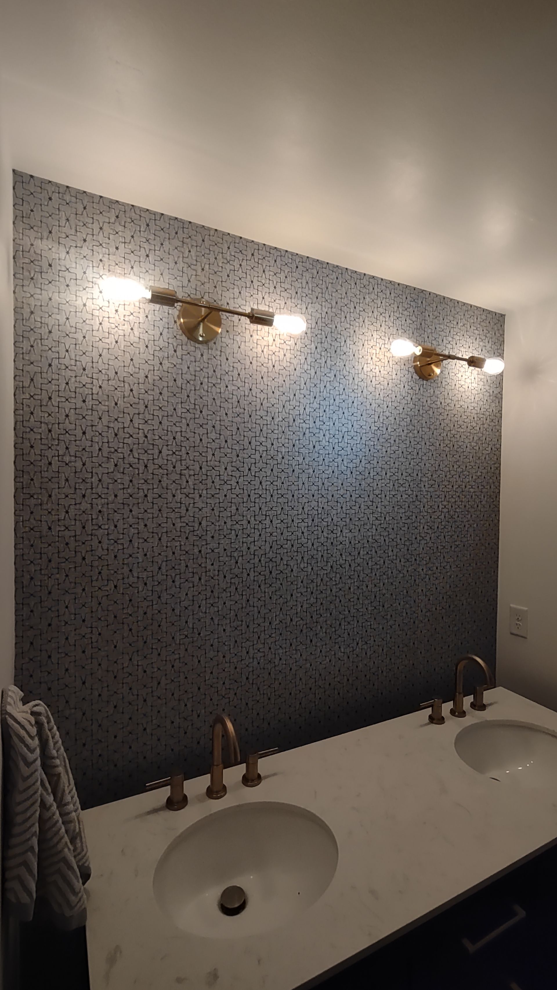 A bathroom vanity with white double sinks, gold faucets, and a patterned tile wall featuring two gold wall-mounted lights.