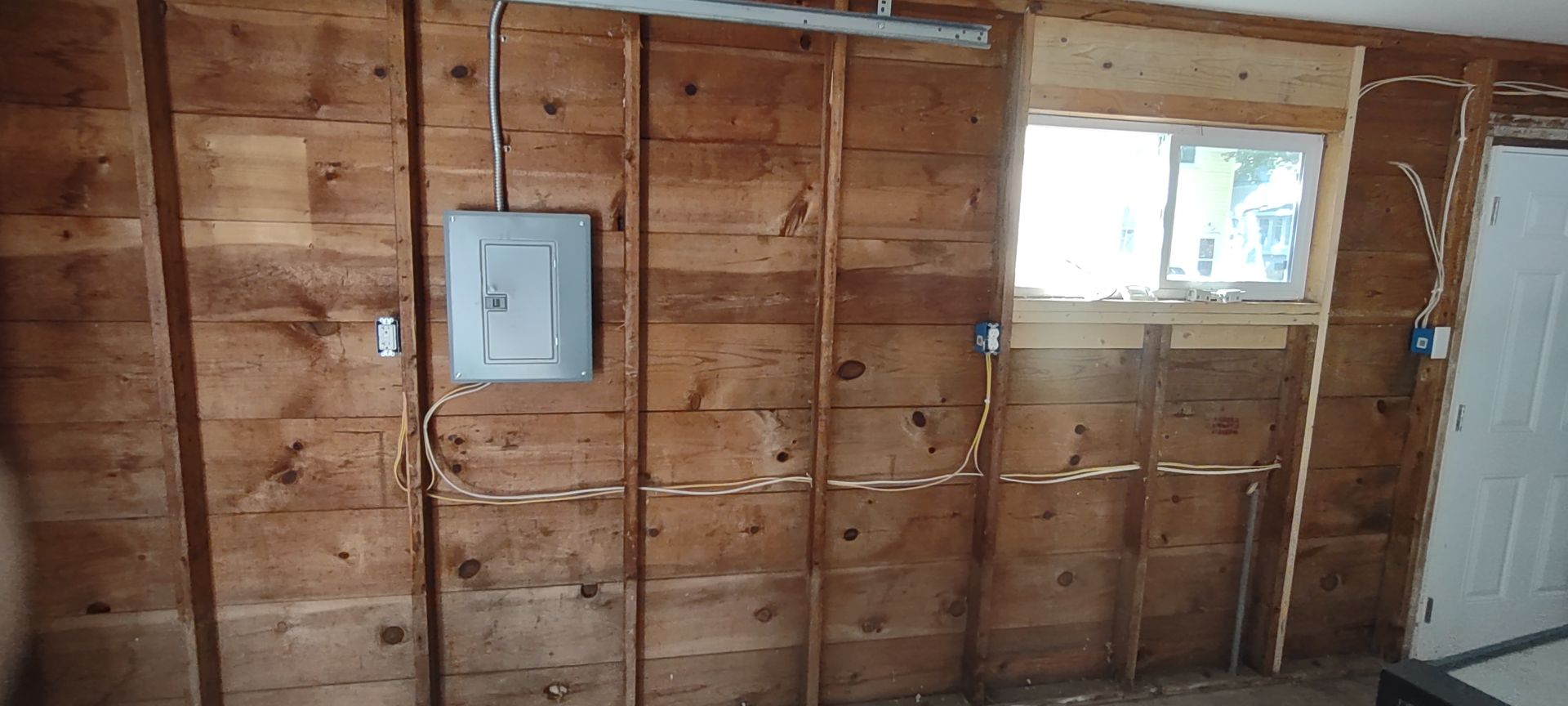A wood-paneled wall inside a garage with an electrical breaker box and exposed wiring connected to outlets.