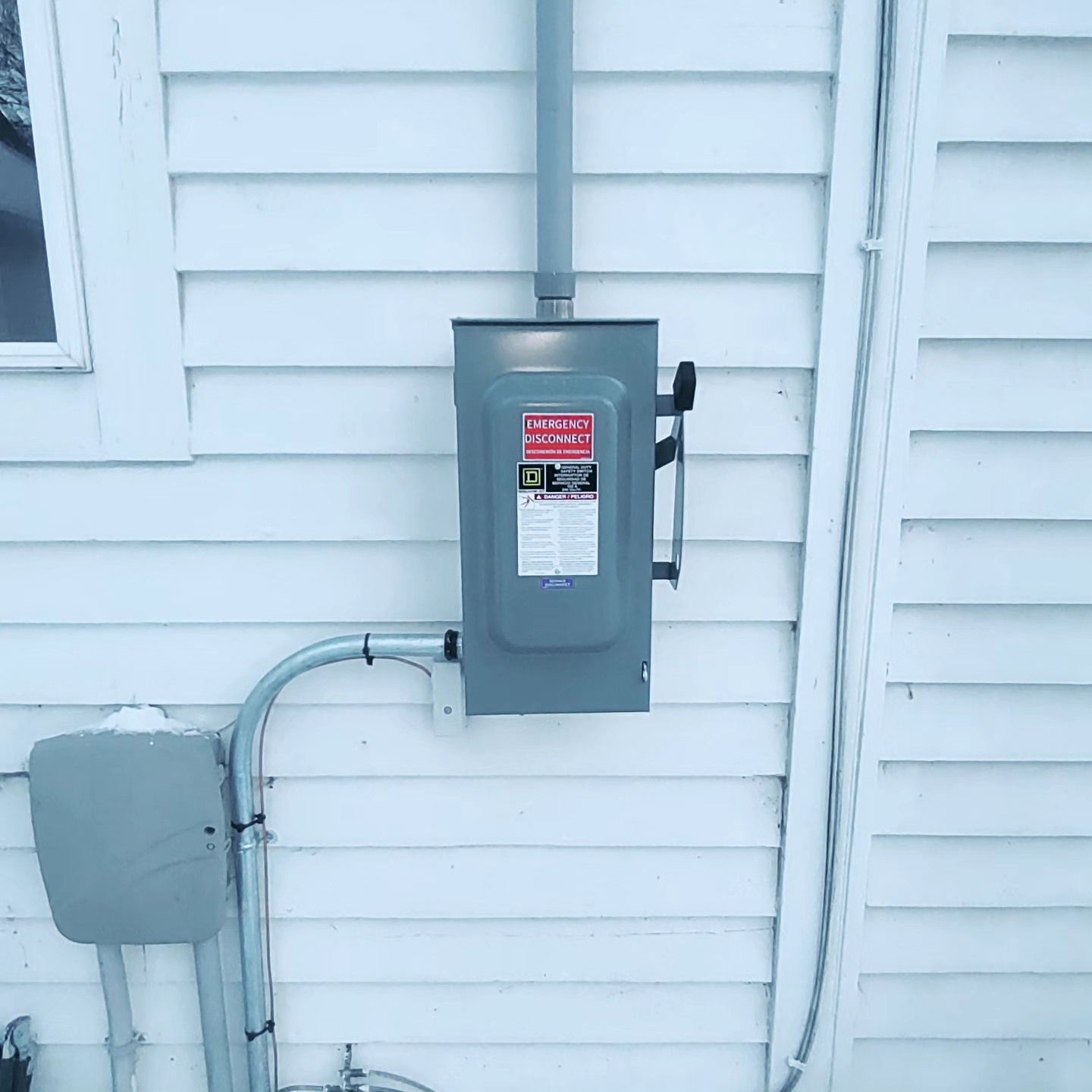 A gray electrical disconnect box mounted on a white exterior wall with conduit running to a smaller junction box below.