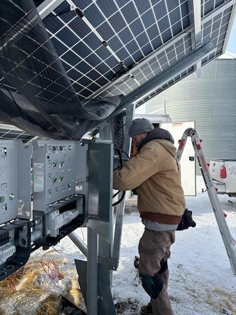 A technician in a brown jacket works on the electrical components of a solar panel array in a snow-covered outdoor area.