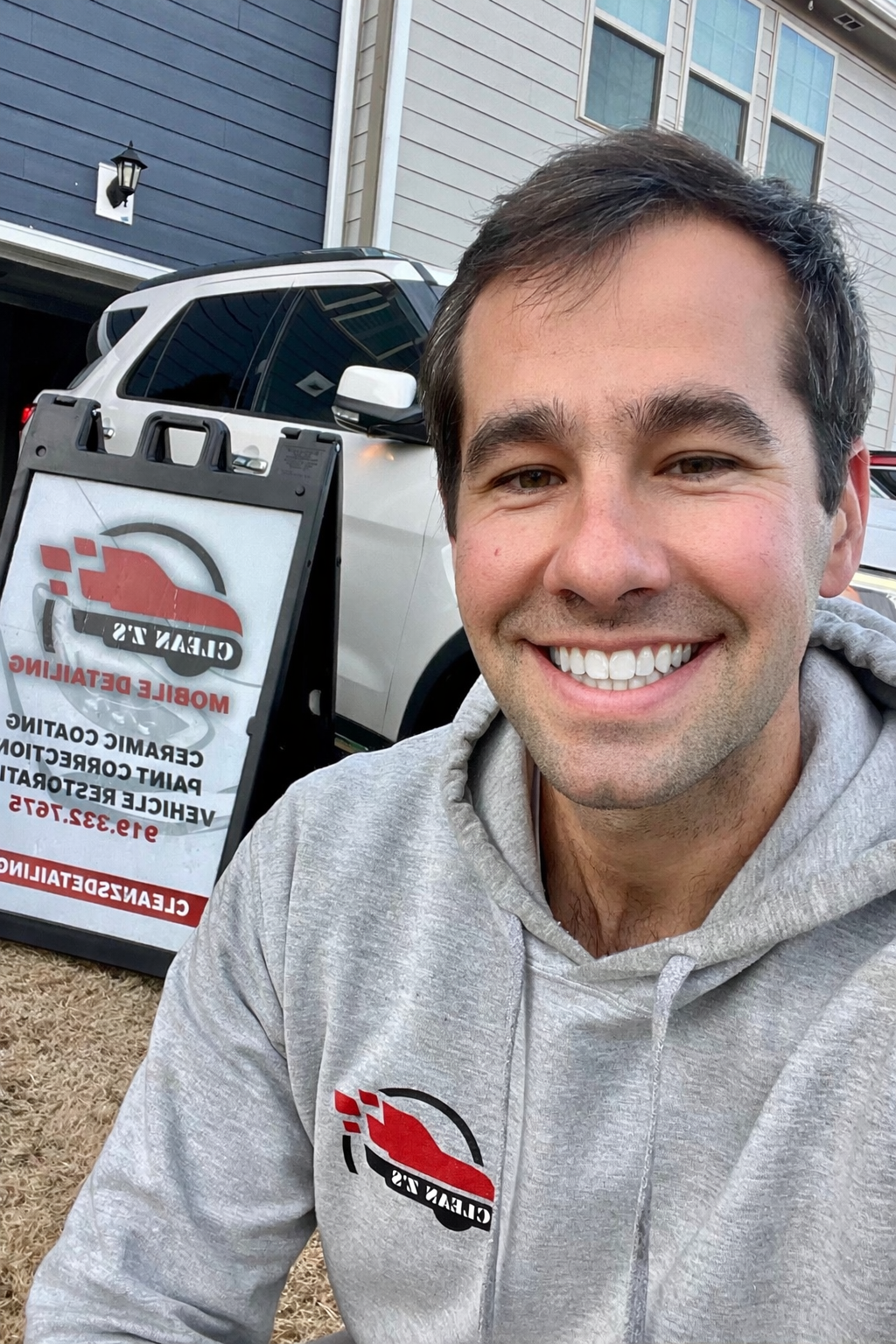 Man smiles in front of a sign and a vehicle, wearing a gray hoodie.