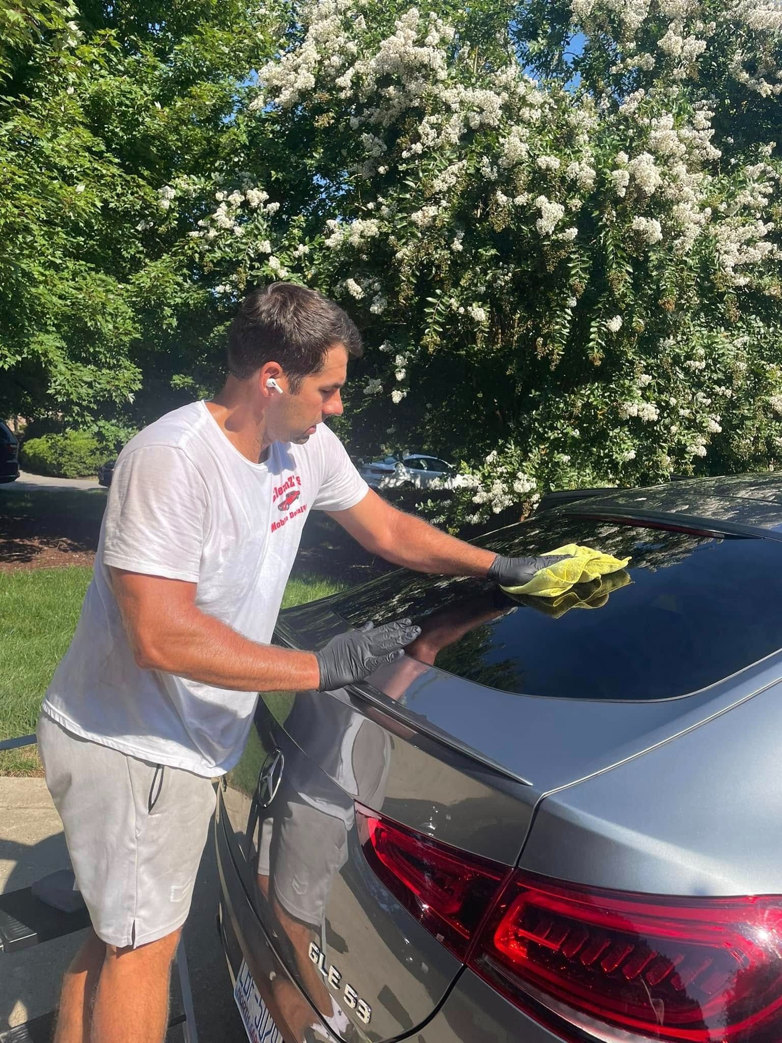 Man washes a silver car in front of a flowering tree, wearing shorts and a white shirt, using a yellow cloth.